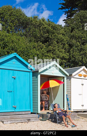 Ältere Frau, die sich im September in Strandhütten sonnte, während ein Mann an einem warmen, sonnigen Tag im Avon Beach, Mudeford, Christchurch, Dorset, Großbritannien, im Schatten saß Stockfoto