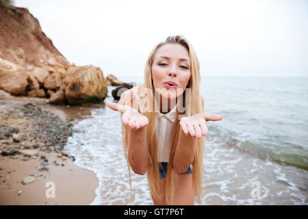 Lächelnden jungen blonden Frau bläst Luft Kuss am felsigen Strand an einem sonnigen Tag Stockfoto