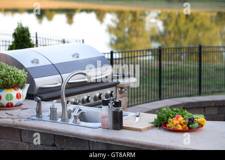 Zubereitung eines gesunden Sommer Menüs in einer Außenküche mit Gasgrill und Waschbecken auf einer Ziegel-Terrasse mit Blick auf einen ruhigen See wi Stockfoto