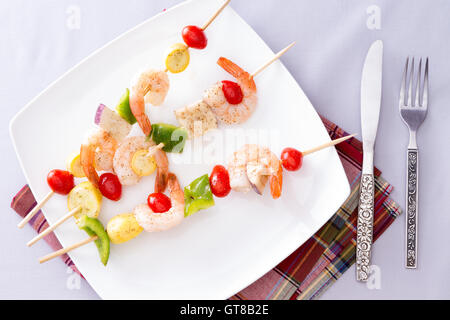 Drei leckere Gourmet-Garnelen Spieße oder Kebabs auf weißem Teller auf einem weißen Tisch mit Servietten und Besteck, eingefangen in hoch ein Stockfoto