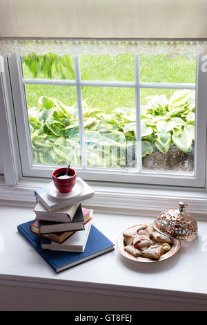 Tasse heißen schwarzen Kaffee auf gestapelten Bücher und Teller mit leckeren Blätterteige auf die Glasfenster für die Lesezeit mit Snacks-Konzept Stockfoto