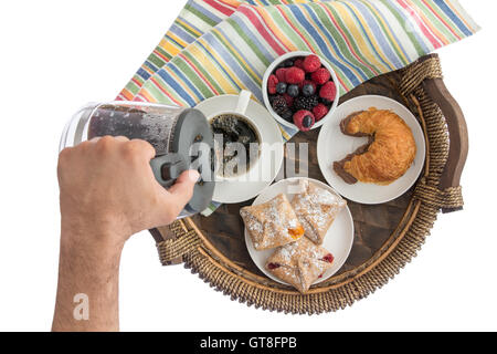 Man gießt frischen Kaffee für sein Frühstück aus einem Glaskrug in eine Tasse auf einem Korb Tablett mit verschiedenen Beeren und Gebäck und ein Stockfoto