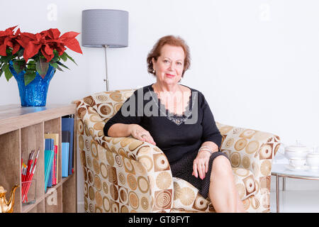 Gnädig elegante senior Frau Entspannung zu Hause in eine bequeme Sessel sitzende lächelnd in die Kamera, festliche rote Weihnachtsstern f Stockfoto