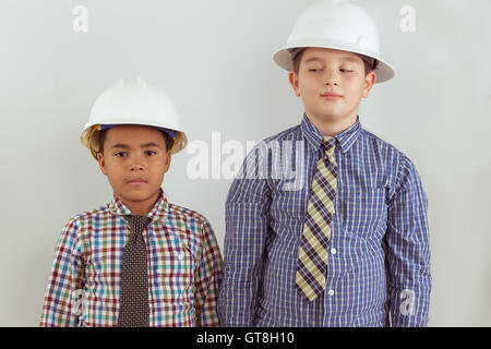 Zwei Rassen junge aufstrebende Tweens Ingenieure gekleidet in Bindungen und Schutzhelme stehen Seite an Seite gegen eine Wand als die zwei frie Stockfoto