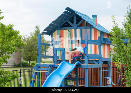 Glückliche kleine Junge in kurzen Hosen sitzen bequem im Sessel auf frisch lackierte persönlichen Spielplatz mit Rutsche und Leitern Stockfoto