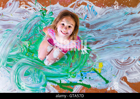 Kreative Farbe bespritzt kleines Mädchen Spaß mit Farben im Zentrum ihrer künstlerischen Moderne Malerei grinsend kniend Stockfoto