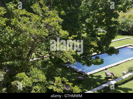 Frau sitzt auf Liegestuhl neben sonnigen Swimmingpool unter grünen Sommer Bäume Stockfoto