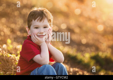 Ein netter Junge Junge lächelt in die Kamera hielt seinen Kopf in seine Hände mit einem funkelnden Bokeh-Hintergrund. Stockfoto