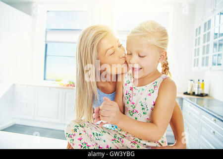 Schöne junge blonde Mutter küssen Tochter in Pferdeschwänze und Kleid, während Sie mit Brotteig spielt Stockfoto