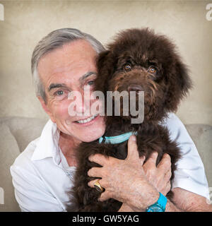 Senior reifer Mann auf Couch halten Haustier Welpen Hund Schokolade Labradoodle. Sie sind Wange an Wange.  Der Mann lächelt. Stockfoto