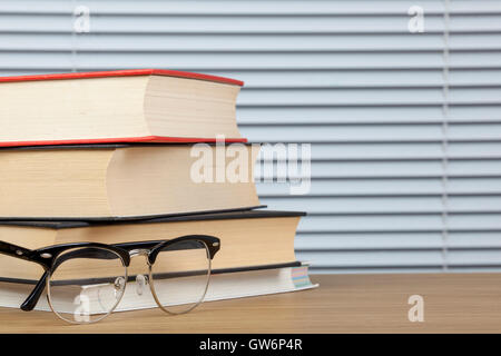 Stapel Rerading Bücher auf einem Tisch mit einer schwarz umrandeten Augen Brille mit Jalousien im Hintergrund Stockfoto