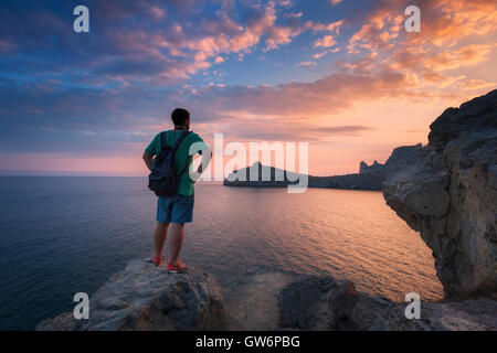 Junge stehender Mann mit Rucksack auf dem Stein am Ufer des Meeres in farbenfrohen Sonnenuntergang Himmel. Wunderschöne Landschaft mit sportlicher Mann, Felsen Stockfoto