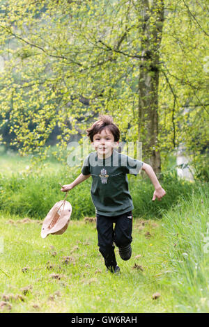 Kaukasier Kind, Junge, 6-8 Jahre alt. Zeigt. In Rasenfläche mit Bäumen hinter, glücklich in Richtung Betrachter holding Floppy hat durch die Schlaufe. Stockfoto