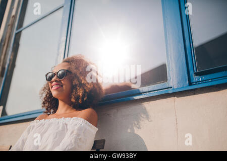 Im Freien Schuss glücklich junge afrikanische Frau im Freien sitzen und Lächeln auf den Lippen. Frau mit Sonnenbrille sitzt vor ihrem Haus.