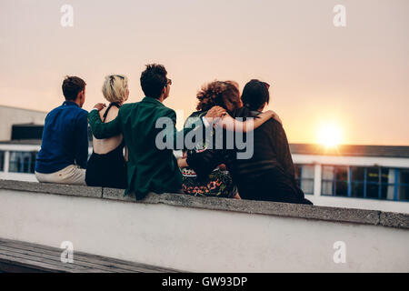 Rückansicht der jungen Männer und Frauen entspannen zusammen auf Dach und Blick auf den Sonnenuntergang. Gemischtrassig Freunde sitzen auf der Terrasse in e Stockfoto