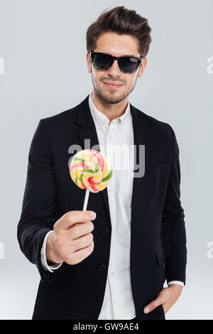 Junger Geschäftsmann in Sonnenbrille stehend mit Hand in der Tasche halten Lutscher auf grauem Hintergrund Stockfoto
