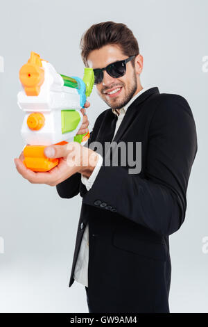 Glücklich lächelnd Geschäftsmann mit Sonnenbrille und Anzug über grauen Hintergrund mit Wasserpistole schießen Stockfoto