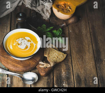 Kürbis Cremesuppe in Schüssel mit Basilikum Blätter, Gewürze und gegrillten Brotscheiben serviert mit frischen Kürbis über alte rustikale Holz b Stockfoto