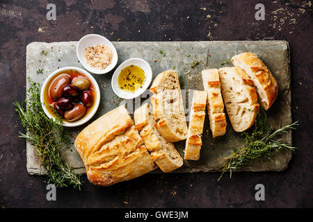 Frisches Ciabatta-Brot in Scheiben geschnitten auf Stein Schiefer Brett mit Oliven, Öl, Salz und Kräutern auf dunklem Hintergrund Stockfoto