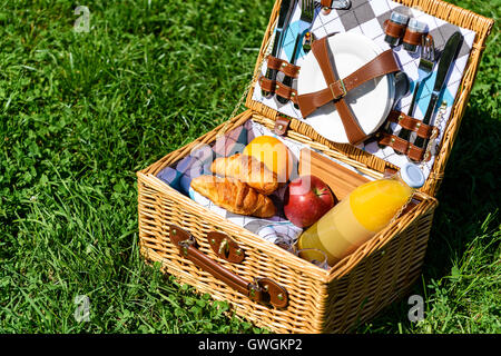 Picknick-Korb mit Flasche Orangensaft, Äpfel, Pfirsiche, Orangen und Croissants auf dem grünen Rasen im Frühjahr Stockfoto