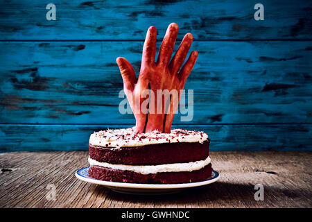 ein roter Samt-Kuchen gekrönt mit einer blutigen Hand für Halloween, auf einer rustikalen Holzoberfläche auf blauem Grund aus Holz Stockfoto