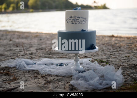 Blauer zweistufiger Hochzeitstorte an einem Sandstrand, mit blauen Steinen dekoriert. Hochzeitstorte auf dem Sand bei Sonnenuntergang Stockfoto