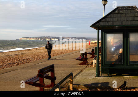 Ältere Dame sitzt im Tierheim am Meer auf einem Winternachmittag, Newhaven im Hintergrund, Seaford, East Sussex, England Stockfoto