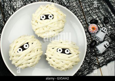 Gruppe von Halloween Mumie Cupcakes auf weißen Teller mit Süßigkeiten auf schwarzem Tuch Hintergrund Stockfoto