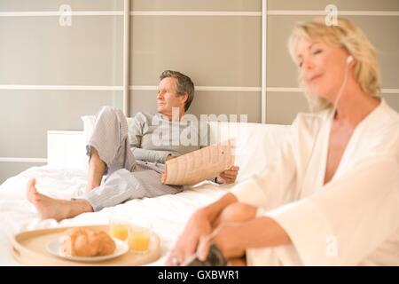 Paar im Bett lesen Zeitung entspannen und Kopfhörer anhören Stockfoto