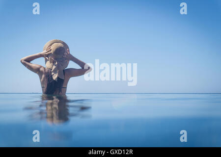 Rückansicht des jungen Frau im Blau des Meeres mit den Händen auf Sonnenhut, Villasimius, Sardinien, Italien Stockfoto