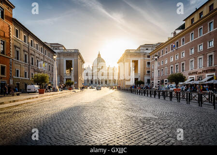 Vatikanische Basilika aus Via della Conciliazione bei Sonnenuntergang mit Sonnenlicht hinter Gebäuden auf Hintergrund Stockfoto