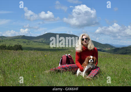 Frau in rot gekleidet, mit ihrem Hund (Cavalier King Charles Spaniel) auf einer Wiese und Zlatibor Berglandschaft im Hintergrund Stockfoto