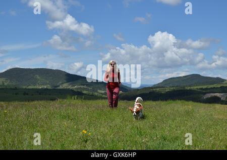 Gekleidete Frau in rot und ihrem Hund (Cavalier King Charles Spaniel) auf einer Bergwiese mit Landschaft im Hintergrund laufen Stockfoto