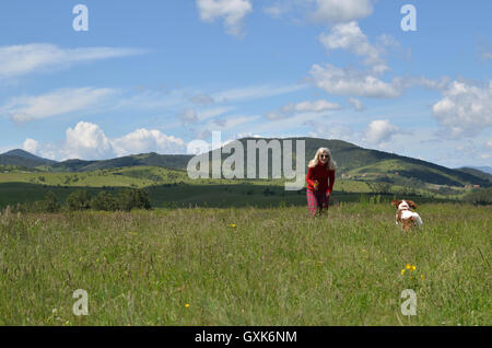 Fröhliche blonde Frau im roten hantierte mit ihrem Hund (Cavalier King Charles Spaniel) auf einer Wiese und wunderbare Landschaft Stockfoto