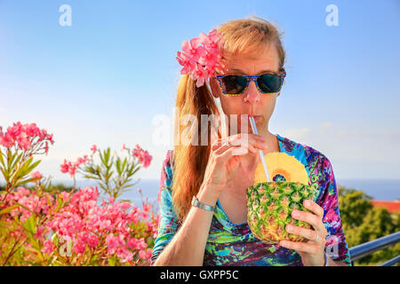 Europäische mittlere gealterte Frau trinkt Kiefer Apfelsaft mit Stroh in der Nähe von rosa oleander Stockfoto
