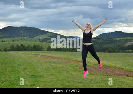 Dame im schwarzen Sportbekleidung mit rosa Sportswear springen auf einer Wiese mit Hügeln und bewölktem Himmel im Hintergrund Stockfoto