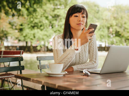 Schuss von attraktiven Asiatin mit Smartphone auf Outdoor-Coffee-Shop. Frauen sitzen im Café-Tisch im Freien mit Handy Stockfoto