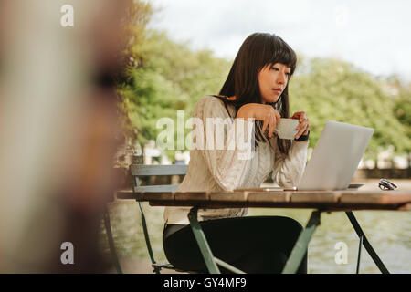 Schuss von schöne junge Frau sitzt an einem Tisch mit einer Tasse Kaffee in der hand mit Blick auf Laptop-Computer. Chinesische Frau, die mit Stockfoto