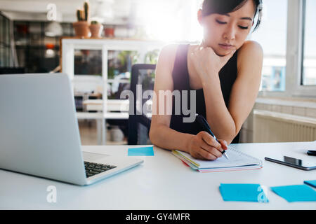 Schuss von asiatischen Geschäftsfrau Schreiben von Notizen auf Notepad. Weibliche Führungskraft sitzt an ihrem Schreibtisch mit Laptop Notizen. Stockfoto