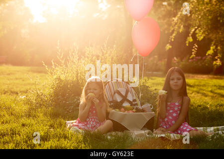 Zwei kleine süße Schwester bei einem Picknick. Mädchen sitzen auf einer Wolldecke und Früchte zu essen. Outdoor-Aktivitäten. Ballons gebunden zu einem Picknickkorb. Stockfoto