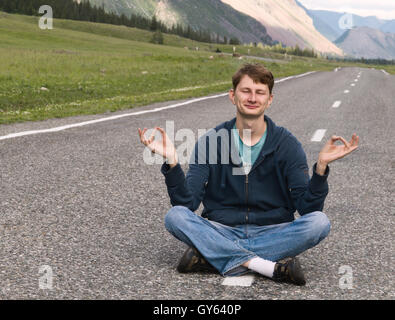 Porträt eines attraktiven jungen kaukasischen Mannes (Kerl, Mann, Person, Modell) sitzen auf der asphaltierten Straße unter Berglandschaft Stockfoto