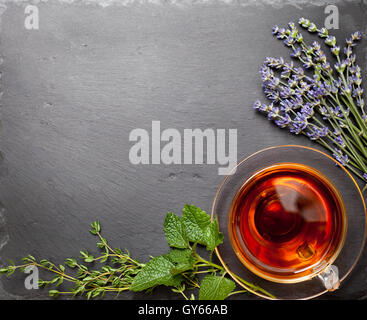 Kräutertee in einem Glas Tasse, verschiedene Arten der Kräuter: Lavendel, Minze, Thymian auf schwarzem Stein (Draufsicht) Stockfoto