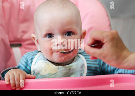 Schöne Baby isst Brei aus Mamas Hand. Er sitzt auf einem rosa Kinder "Stuhl. Stockfoto