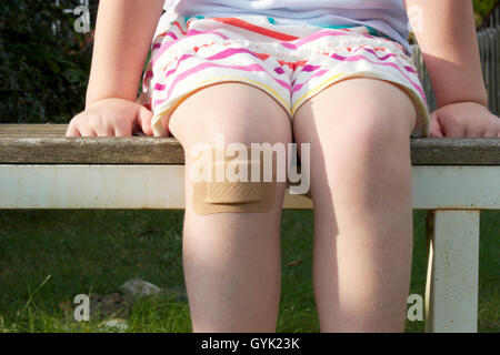 Ein kleines Kind auf eine Gartenbank mit einem großen Pflaster auf ihrem Knie sitzt. Stockfoto