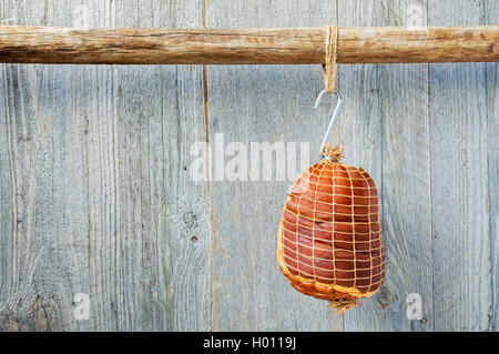Geräuchert ohne Knochen Schweinefleisch Schinken Hock eingewickelt in Netzen hängen an einem Haken aus einem Holzstab mit einem verwitterten Holz Hintergrund Stockfoto