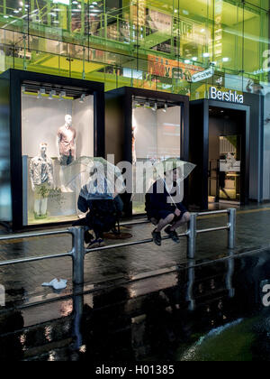 Zwei jungen mit passenden Sonnenschirmen sitzen Rücken an Rücken in den Regen draußen ein Modegeschäft, Shibuya, Tokyo Japan Stockfoto