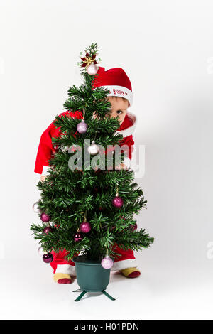 Baby Boy verkleidet als Weihnachtsmann versteckt sich hinter Weihnachtsbaum. weißen Hintergrund. Stockfoto