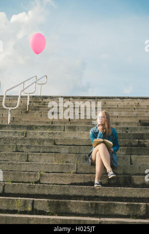 Nachdenkliche junge Schülerin sitzt auf Außentreppen mit Buch. Ballon auf Hintergrund Stockfoto