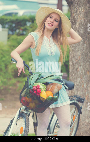 Attraktive blonde Frau mit Strohhut im Park stehen und posiert neben Fahrrad mit Korb voller Lebensmittel. Post-Geschäftsprozeß Stockfoto