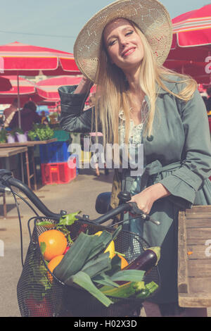 Attraktive blonde Frau mit Strohhut und Fahrrad auf dem Marktplatz. Post mit Vintage-Filter verarbeitet. Stockfoto
